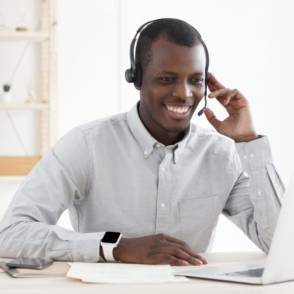 Smiling african customer support service operator with hands-free headset working in office, using laptop